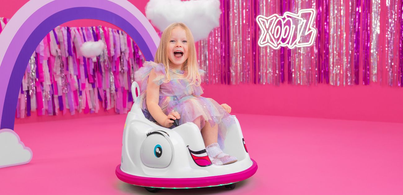 A young girl with blonde hair riding a unicorn bumper car. The background is pink with a purple rainbow and sparkly tinsel.