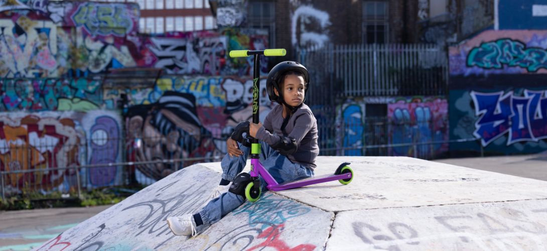 A photo of a young girl sat on a skate ramp holding a purple and green stunt scooter