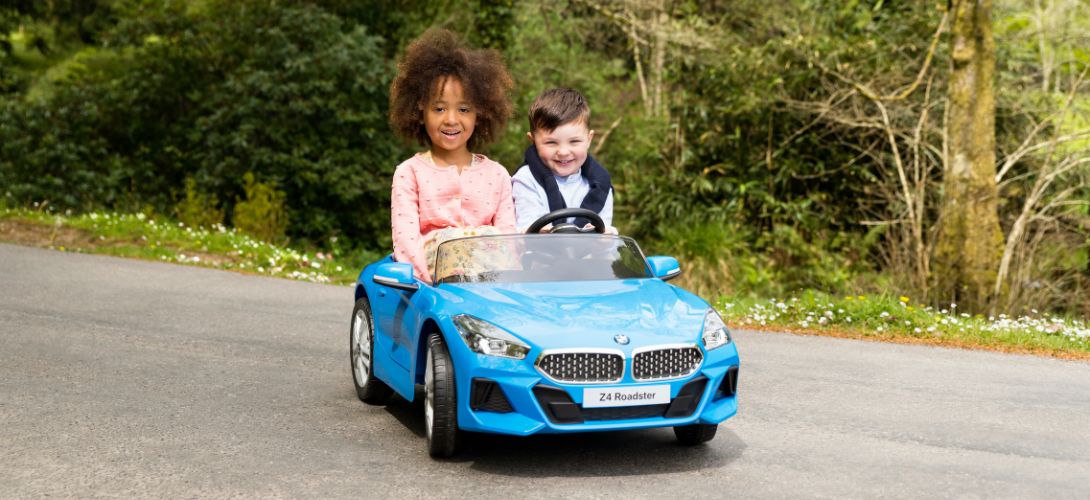 A photo of a boy and girl sat in a blue BMW ride on car with a wooded area behind her
