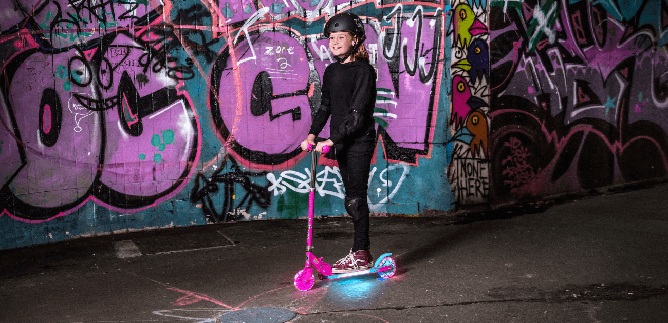 Girl riding a Xootz LED scooter near a graffiti wall, highlighting fun outdoor activities for summer.