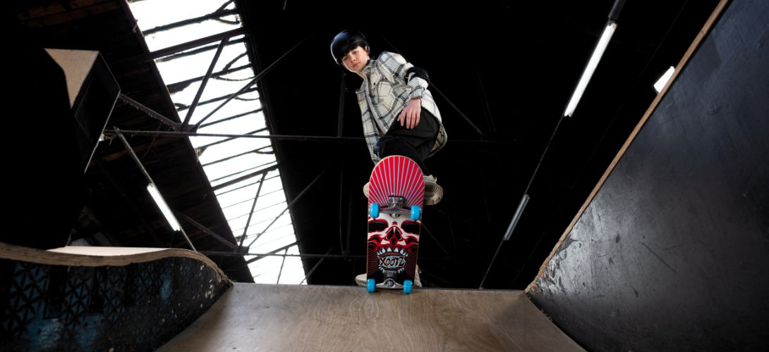 A photo of the underneath of a skateboard as a boy performs a trick on a ramp. The deck has a red skull print design with blue wheels