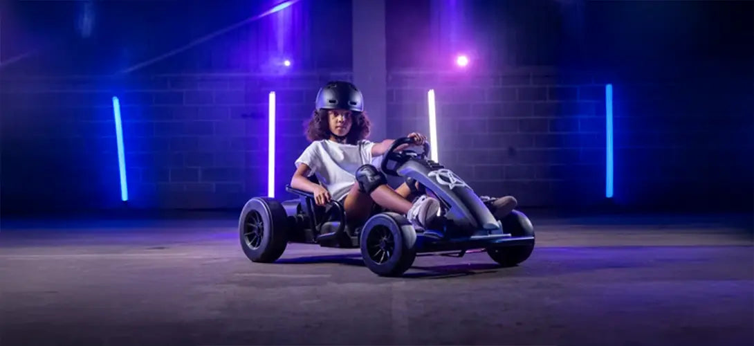 A photo of a girl riding a black and green go kart. She is in a dark warehouse which is lit with white and green lights